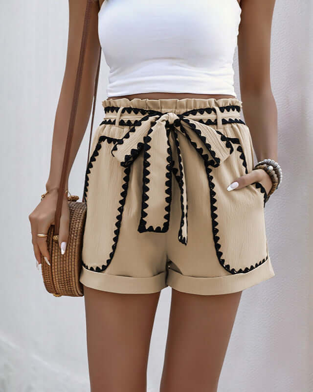 Front view of beige high-waisted shorts with black lace trim and a bow belt, paired with a white crop top and accessorized with a woven bag.
