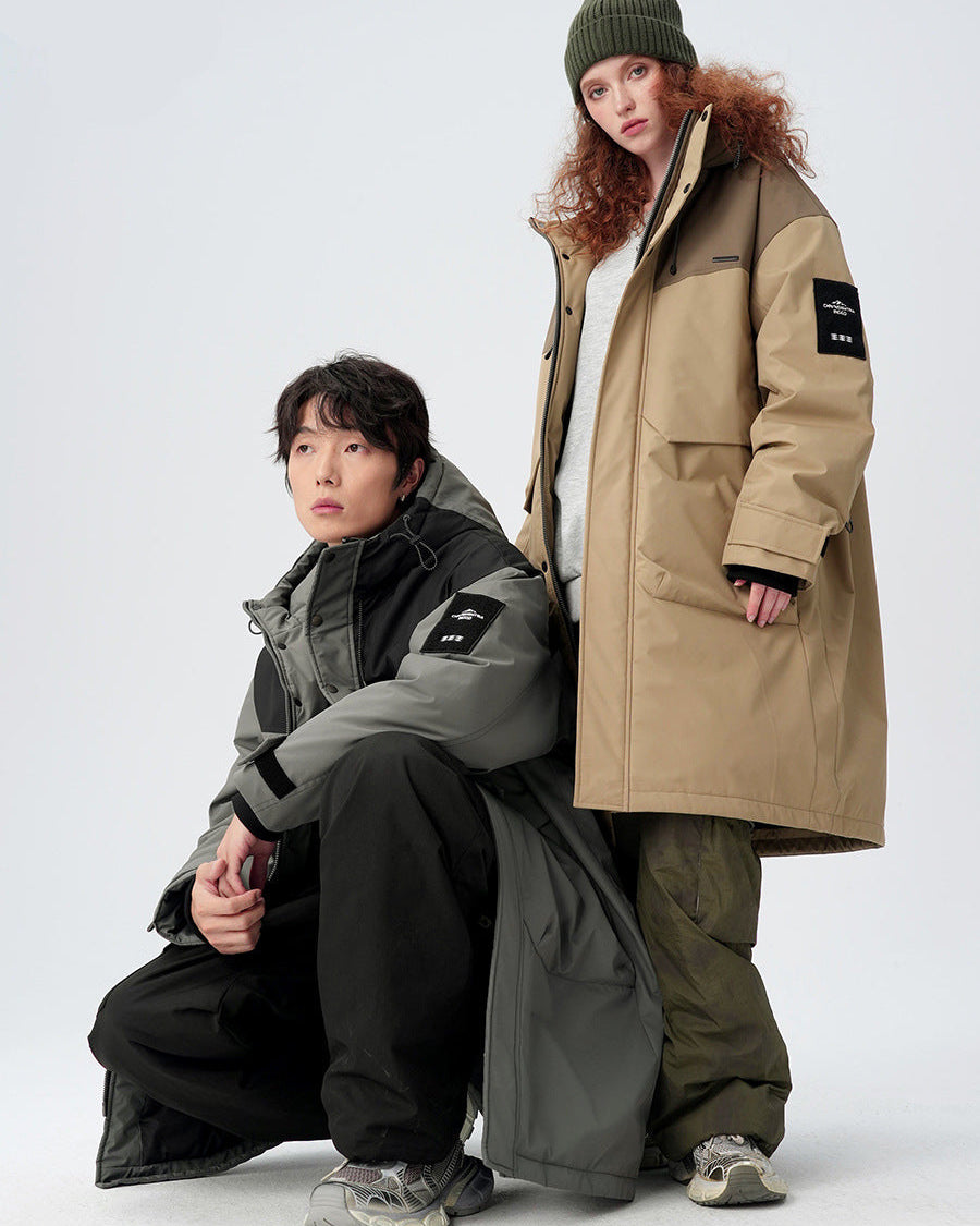 A woman in olive beanie stands while man kneels, both wearing technical parkas in contrasting colors against white background