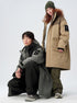 A woman in olive beanie stands while man kneels, both wearing technical parkas in contrasting colors against white background