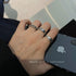 A person wearing three silver rings on their fingers - a chain-link, Roman numeral, and twisted band ring - while holding a dark iPhone