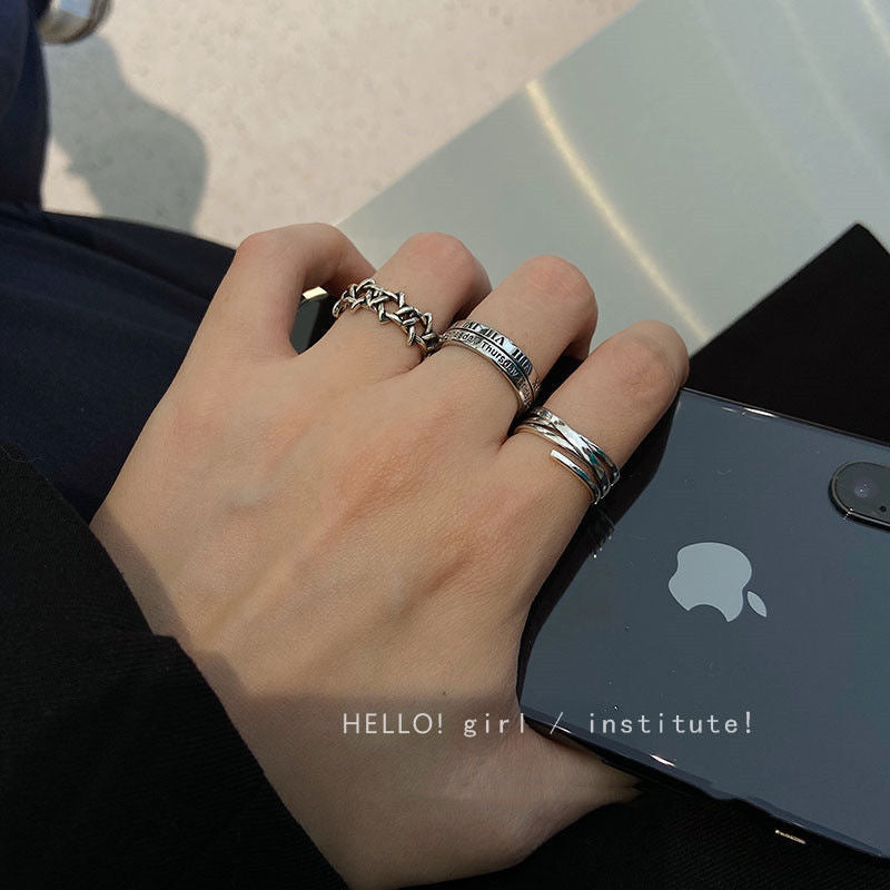 A person wearing three silver rings on their fingers - a chain-link, Roman numeral, and twisted band ring - while holding a dark iPhone