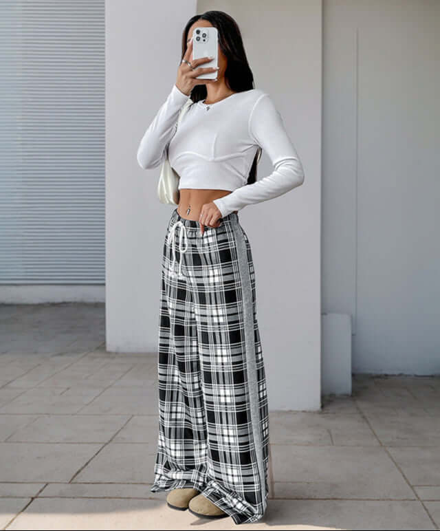Woman taking a mirror selfie wearing black and white plaid lounge pants with a white long-sleeve crop top and a shoulder bag.