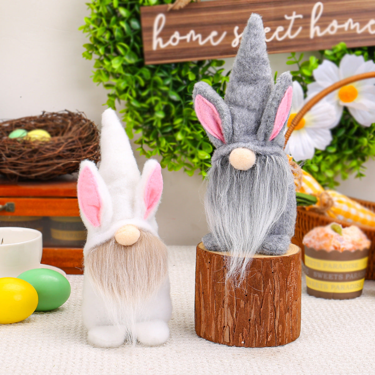 White and gray Easter gnome plush toys, with pink inner ears, posed on a wooden block against a festive "Home Sweet Home" sign.