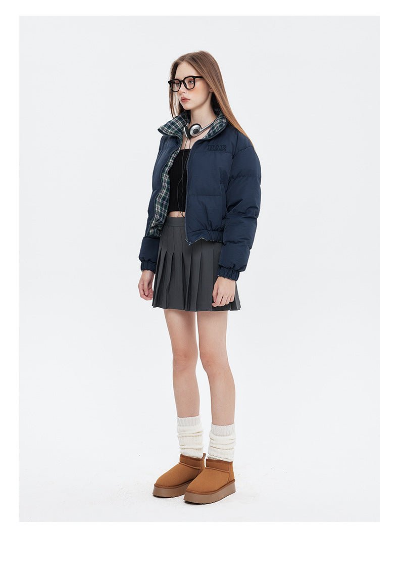 A woman stands in a navy puffer jacket with a plaid lining, a black crop top, a gray pleated skirt, and brown platform boots