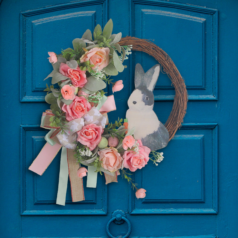 A grapevine wreath adorned with pink roses, green leaves, and a bunny figure, displayed on a blue door.