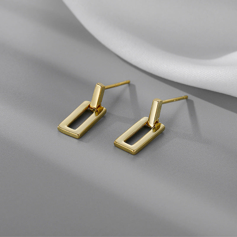 A pair of gold rectangular drop earrings with a sleek, modern design, placed on a gray surface with white fabric in the background.