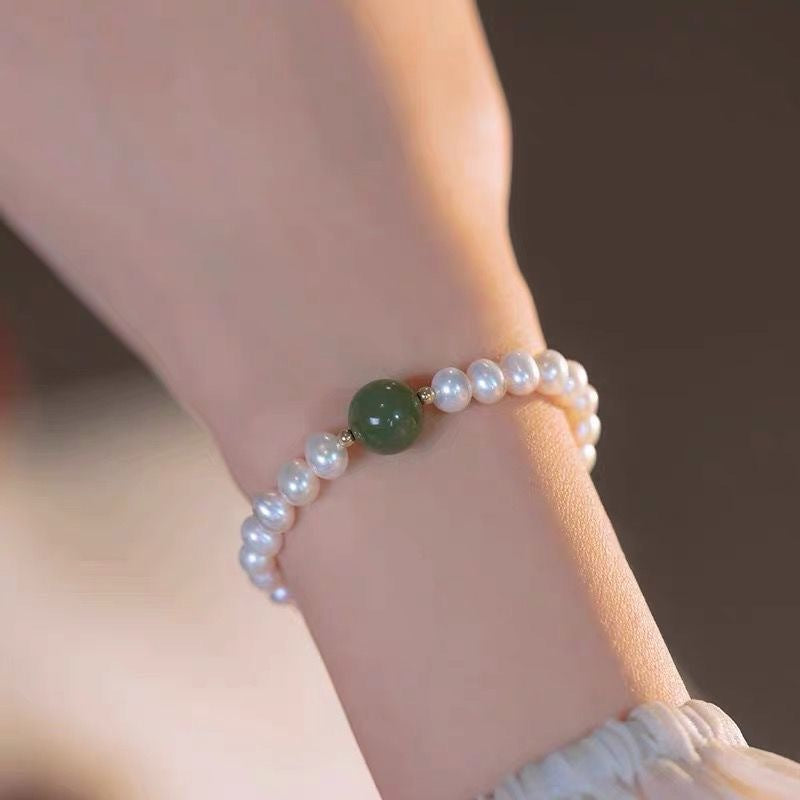 Close-up of pearl bracelet on wrist, showcasing single jade green bead between white pearls against neutral background