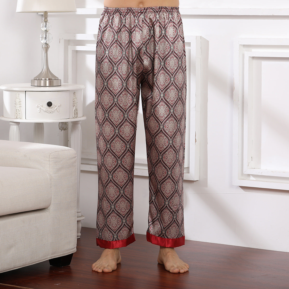 Model wearing burgundy silk pajama pants with ornate diamond pattern and red trim at cuffs, standing barefoot on wooden floor beside white nightstand