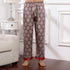 Model wearing burgundy silk pajama pants with ornate diamond pattern and red trim at cuffs, standing barefoot on wooden floor beside white nightstand