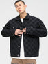 A model is wearing a black checkered denim jacket over white t-shirt and tan pants, posing against white background