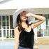 Woman in a white wide-brimmed sun hat shielding her face from the sun, wearing a black velvet camisole and denim jeans.