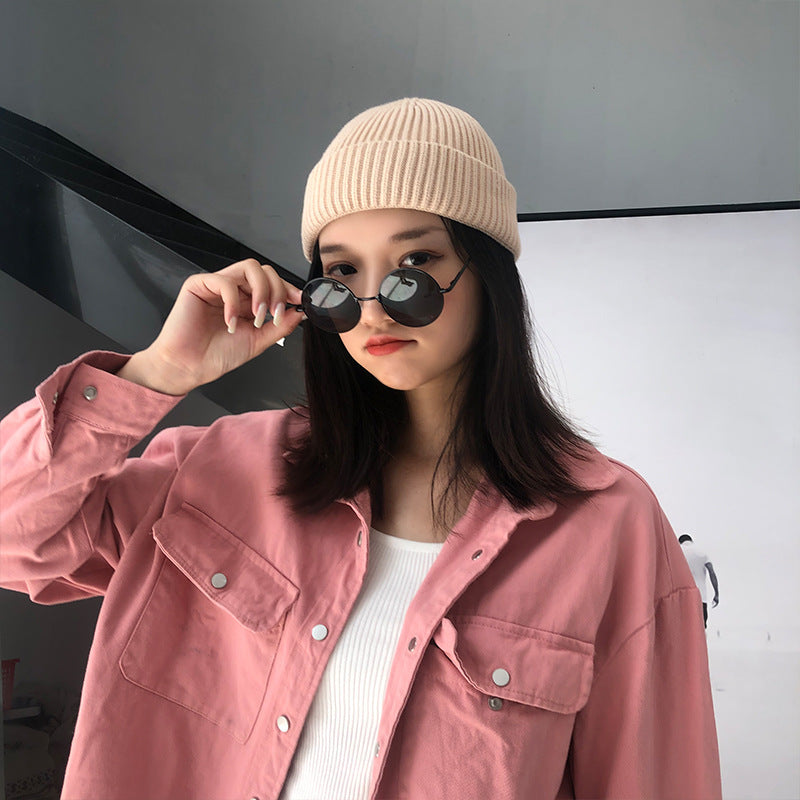 A woman wearing a cream ribbed beanie, round sunglasses and pink jacket, touching frames while posing against white and black backdrop