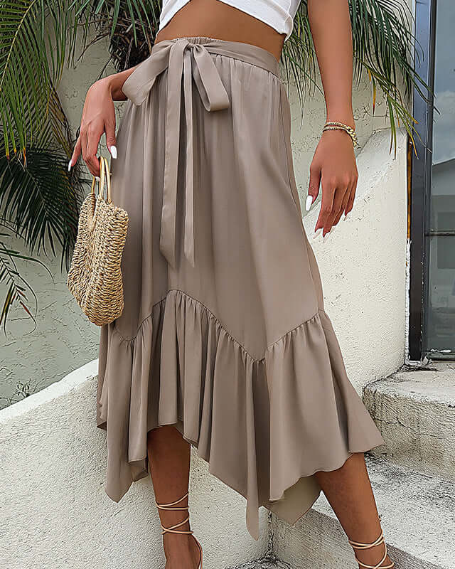 Woman in a khaki midi skirt with a high waist tie and ruffle hem, accessorized with a woven bag and gold jewelry.