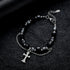 A black gemstone bracelet with silver beads and a cross pendant displayed on a textured black surface, featuring a double chain for an edgy look.