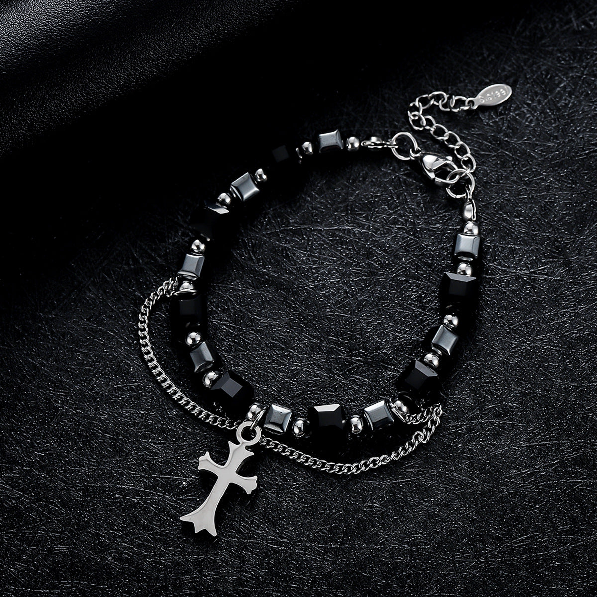 A black gemstone bracelet with silver beads and a cross pendant displayed on a textured black surface, featuring a double chain for an edgy look.