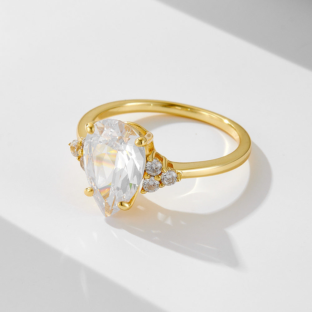 A gold engagement ring with pear-shaped diamond center stone and accent diamonds on each side, displayed on white surface casting soft shadow
