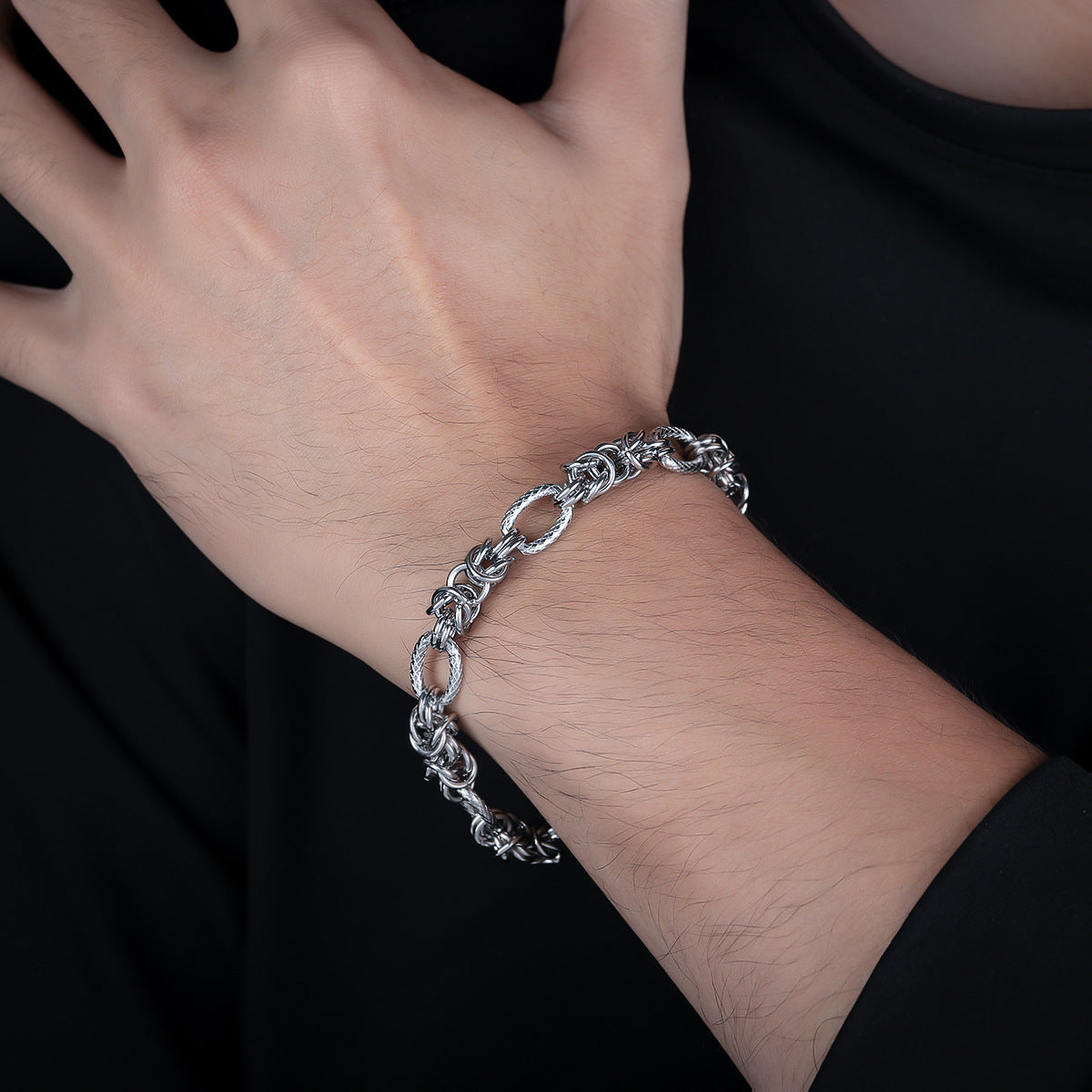 A man’s hand resting on his chest, wearing a silver rope-link bracelet with interwoven details, paired with a black shirt.