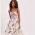 A model with curly brown hair is wearing a white spaghetti strap dress with pink rose and green leaf print, shown from chest up against white background