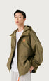 A man wearing olive green zip-up windbreaker with orange bicycle logo detail, styled with white t-shirt against neutral background