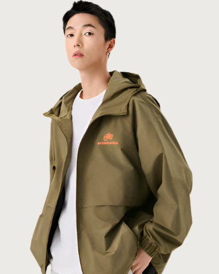 A man wearing olive green zip-up windbreaker with orange bicycle logo detail, styled with white t-shirt against neutral background