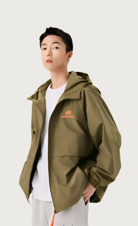 A man wearing olive green zip-up windbreaker with orange bicycle logo detail, styled with white t-shirt against neutral background