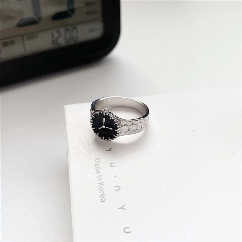 A silver watch-design ring with black face and clock hands displayed on white paper with Korean text in background