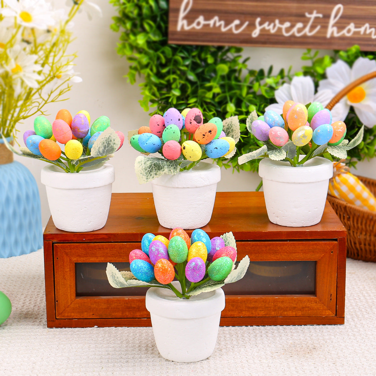 Four white flower pots, each holding a bouquet of artificial speckled Easter eggs in pastel and vibrant shades, displayed on a wooden box with floral decor.