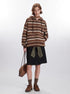 A woman wearing striped brown zip-up hoodie with textured pattern, black denim skirt and olive knit belt, styled with brown socks and shoes against white background