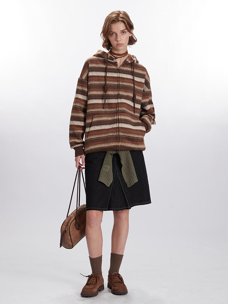 A woman wearing striped brown zip-up hoodie with textured pattern, black denim skirt and olive knit belt, styled with brown socks and shoes against white background