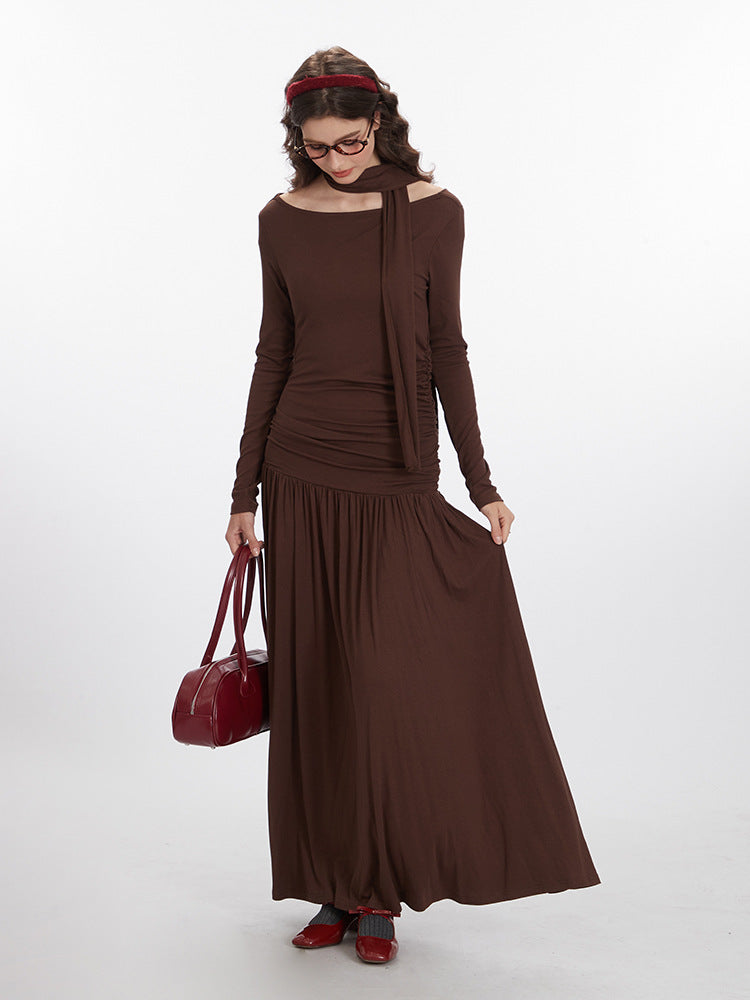 Side view of brown jersey maxi dress highlighting dropped waist silhouette, ruched bodice detail, and attached neck scarf, paired with burgundy accessories
