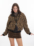 A woman wearing a leopard print bomber jacket with black trim over a brown turtleneck paired with a corduroy mini skirt, with arms outstretched against white background