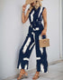A women wearing navy wide-leg jumpsuit with a white abstract print, cinched waist, and sleeveless cut, accessorized with a straw hat and woven bag.