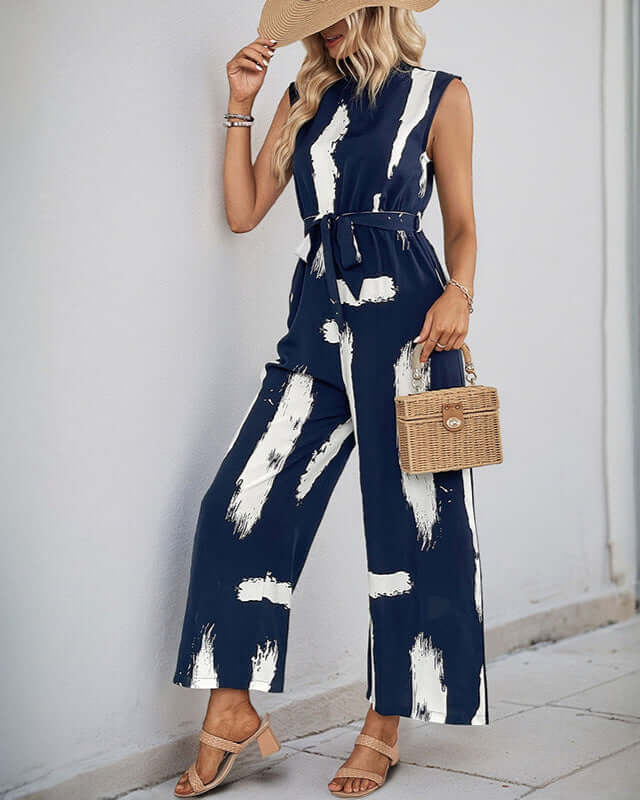 A women wearing navy wide-leg jumpsuit with a white abstract print, cinched waist, and sleeveless cut, accessorized with a straw hat and woven bag.