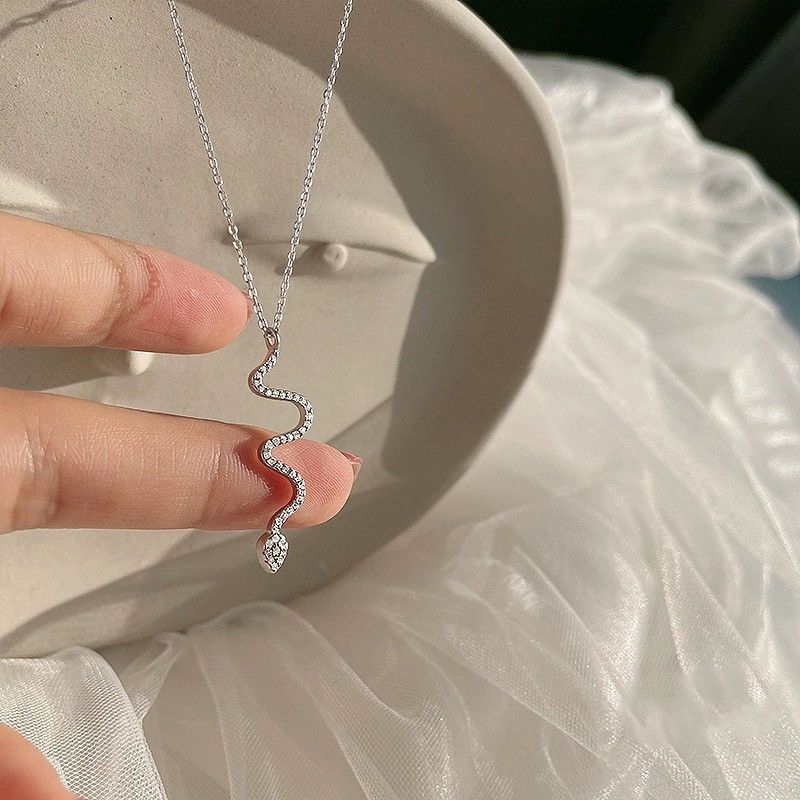 A person holding a delicate silver serpent necklace with diamond-like stones against neutral fabric and ceramic background