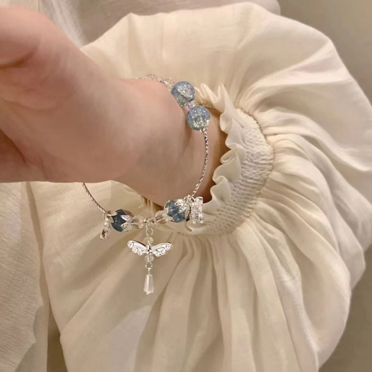 A silver and blue crystal bracelet with butterfly charms worn on wrist with elegant white ruffle sleeve details