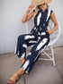 Woman sitting in a navy sleeveless jumpsuit with a white abstract print, styled with a straw hat and bracelets.