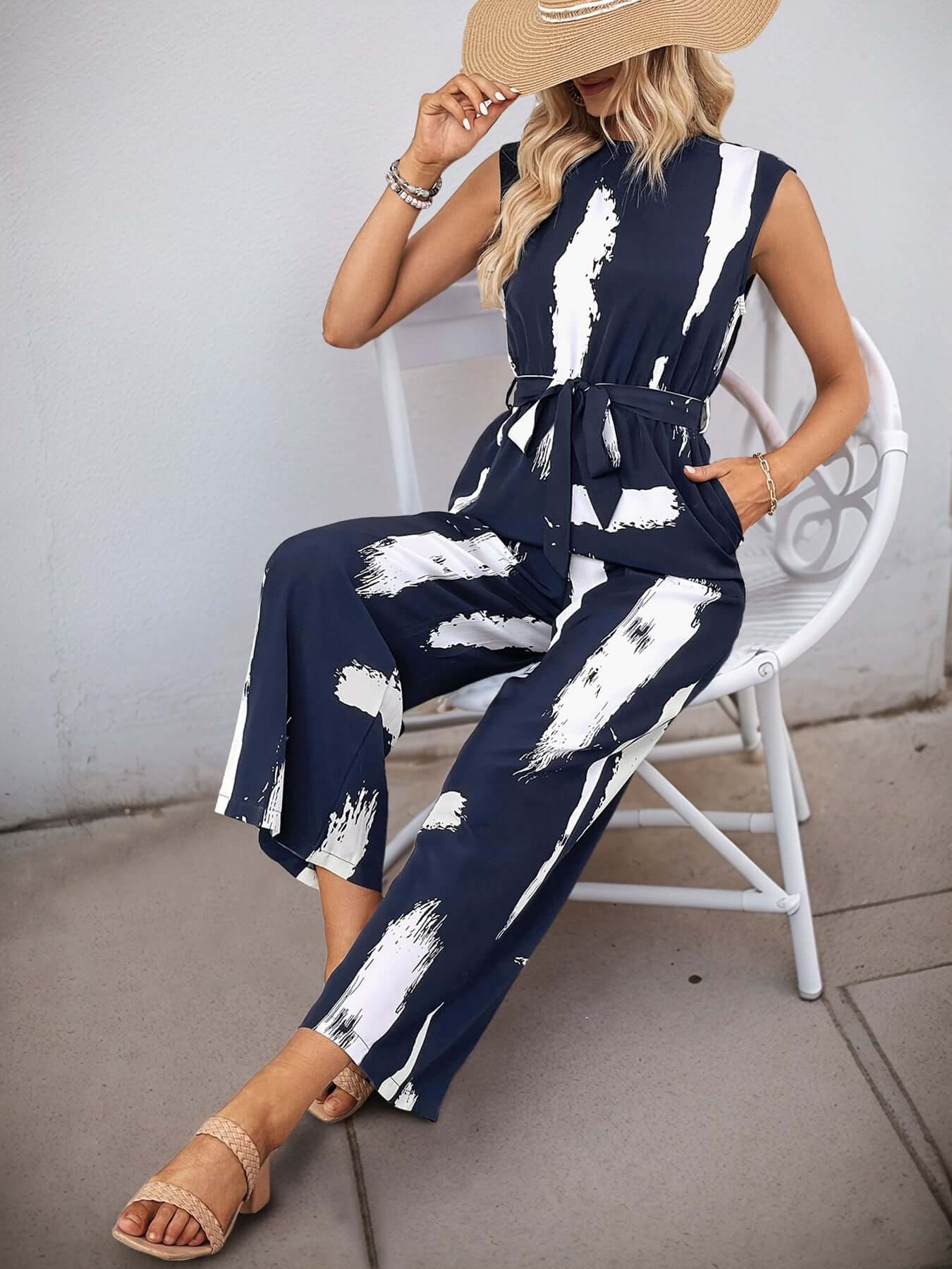 Woman sitting in a navy sleeveless jumpsuit with a white abstract print, styled with a straw hat and bracelets.