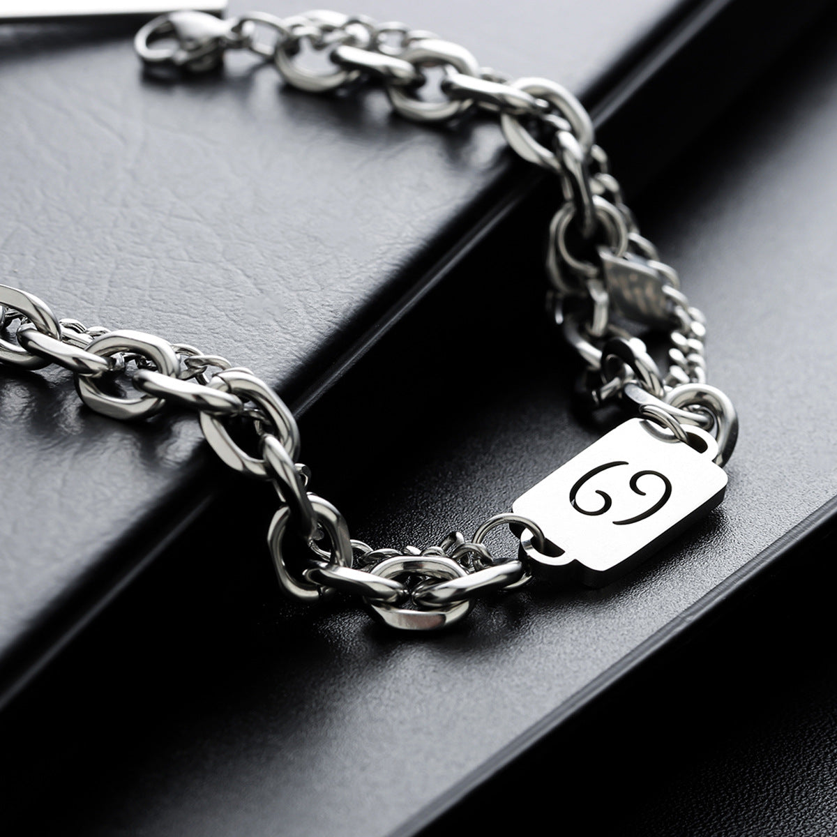 Stainless steel zodiac bracelet with a Cancer symbol charm, displayed on a black leather surface for a sleek, modern look.