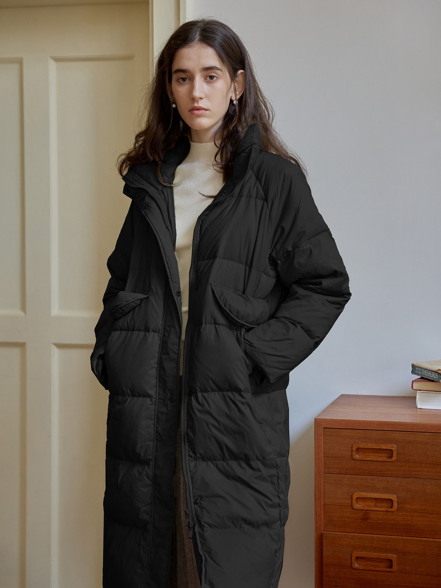 Black maxi puffer coat with high collar and utility pockets, styled with cream turtleneck, minimalist interior setting