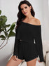 Woman in black knit romper with off-shoulder neckline and long sleeves, sitting on white couch near plant