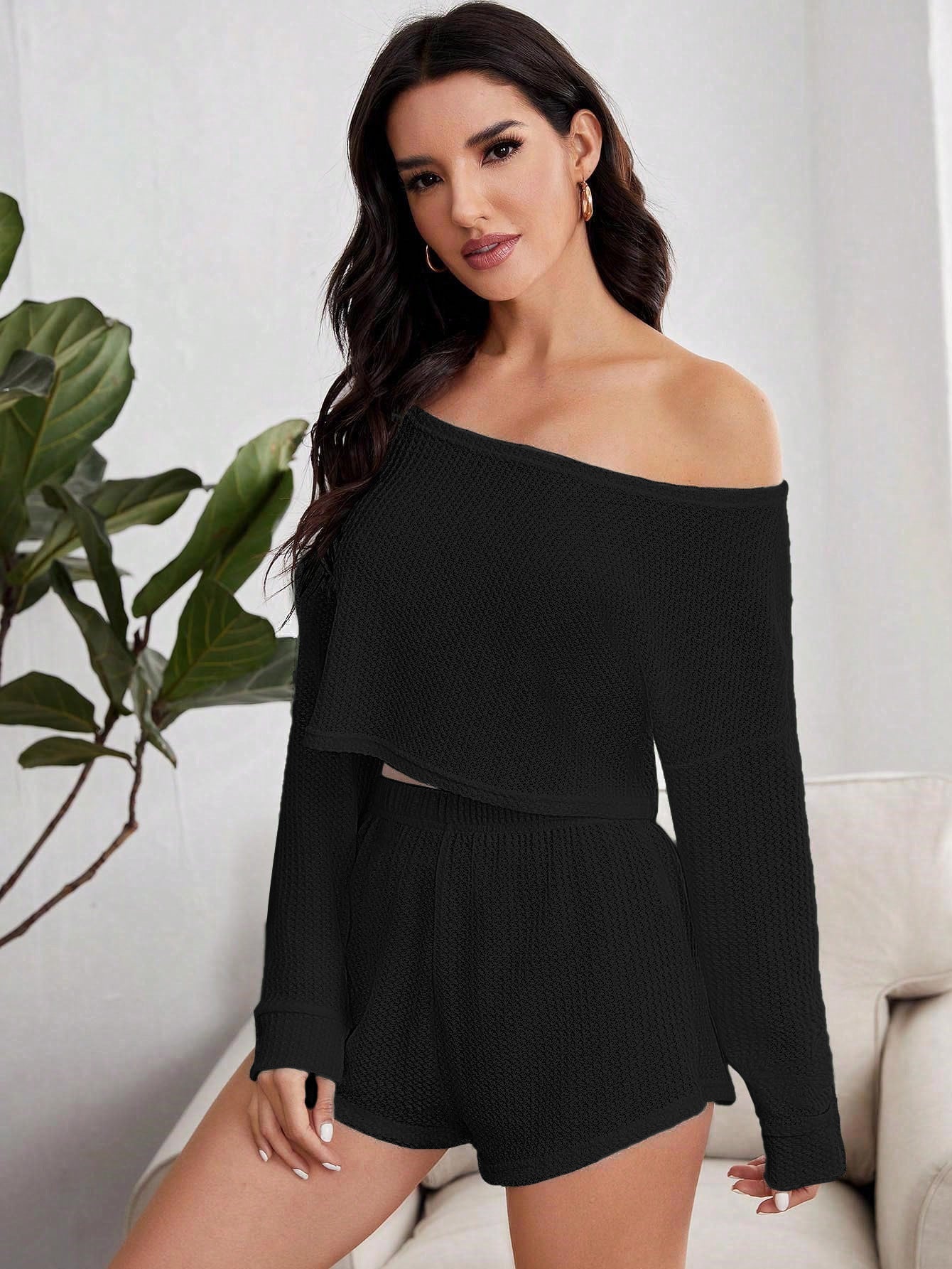 Woman in black knit romper with off-shoulder neckline and long sleeves, sitting on white couch near plant
