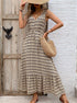 Woman in a beige and black boho maxi dress, holding a woven bag while standing by a rustic wooden door, wearing strappy sandals.