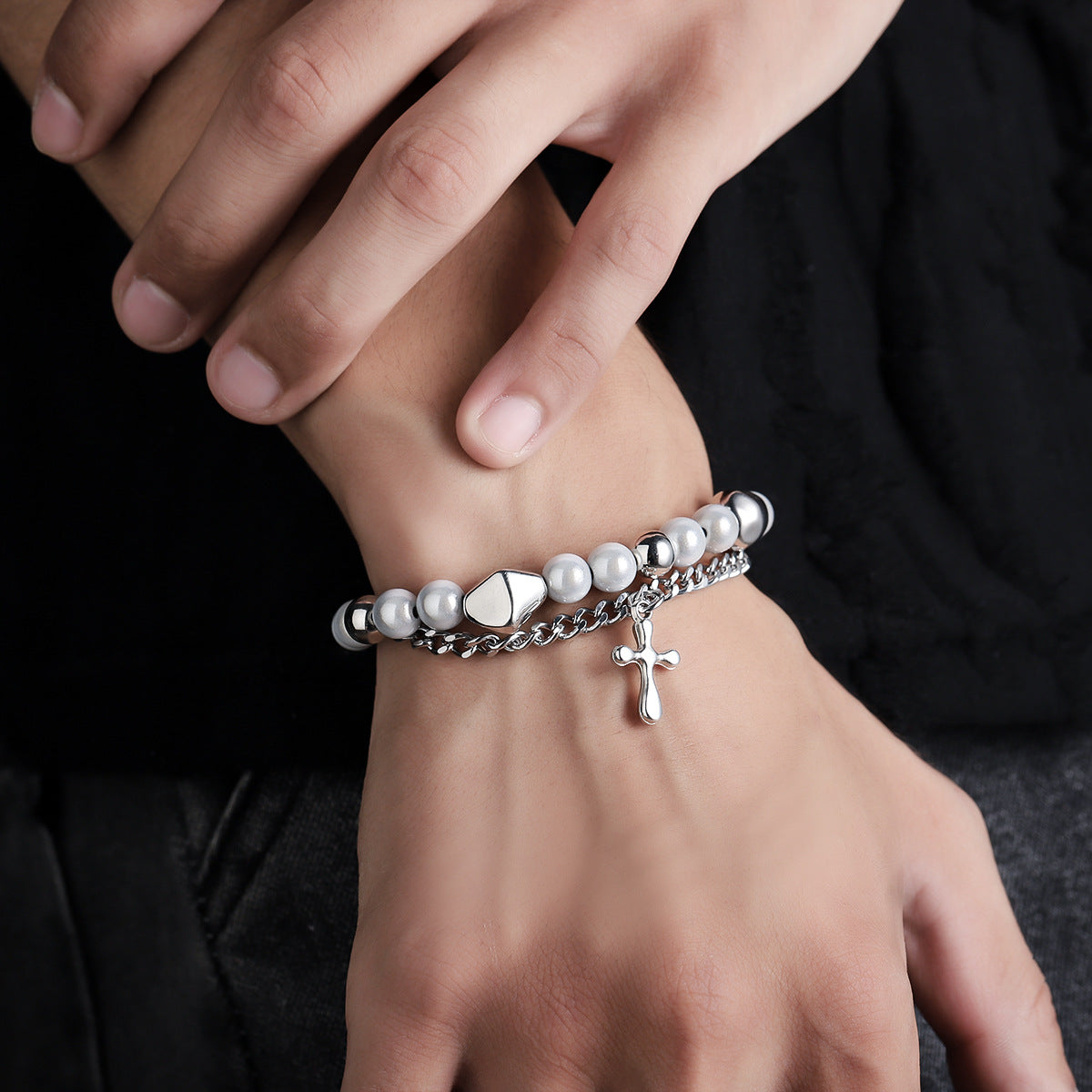 A close-up of a person wearing a silver and white beaded bracelet with a dangling cross charm, styled with a layered chain design, against a black outfit.