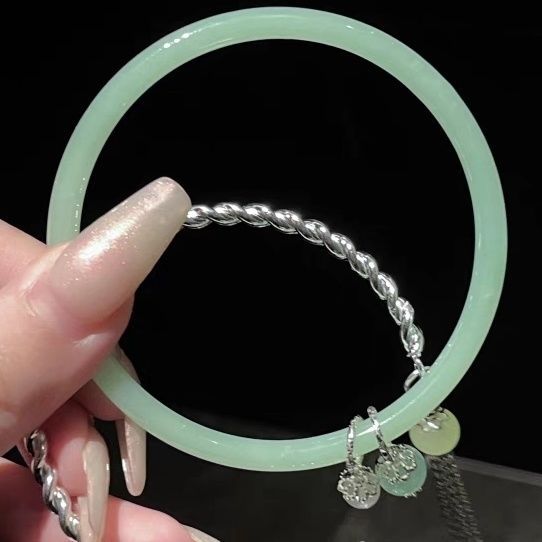 A light jade bangle with silver twisted chain and decorative charms displayed against black background