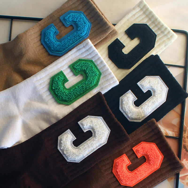 A close-up of several varsity-style crew socks in various colors, each featuring a bold "C" patch in textured fabric