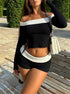 Woman posing with raised hand in black crop top with white trim, matching shorts, white handbag and necklace