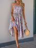  A woman in a floral print dress with a high slit, holding up the skirt slightly while posing. She accessorizes with a woven handbag and layered bracelets.