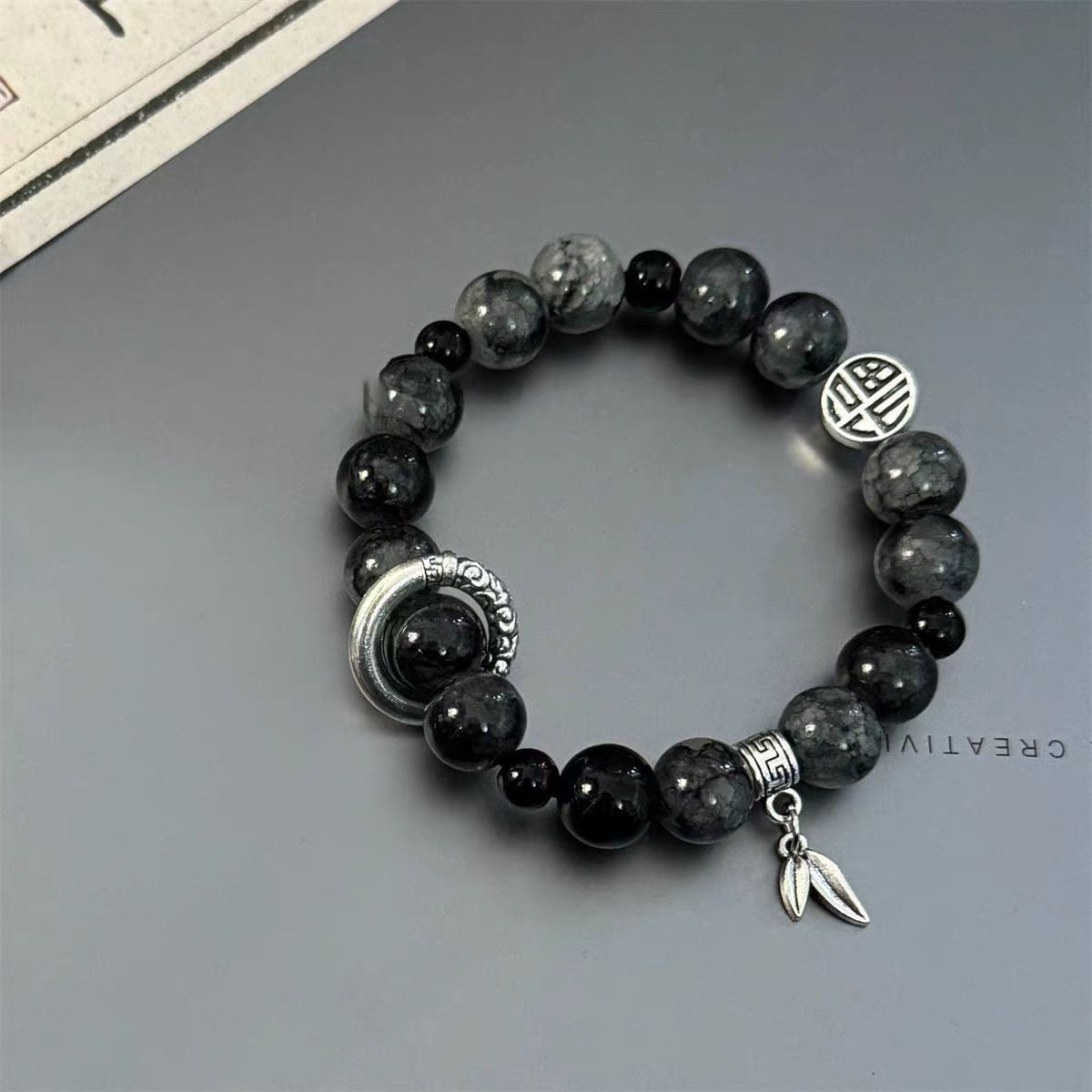 Dark stone bracelet with silver moon, geometric bead and leaf charm on gray background