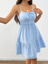 A woman is wearing a baby blue cotton sundress with adjustable straps and gathered tiers, posing against a white wall with wooden flooring