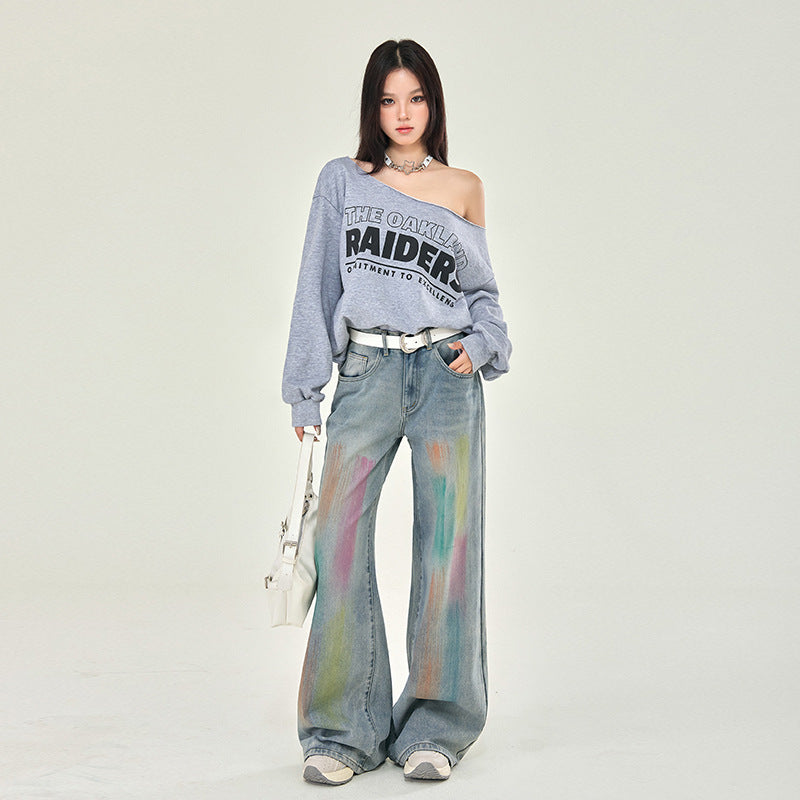 A model wearing an off-shoulder Raiders sweatshirt, wide-leg jeans with pastel tie-dye effect, white belt, and sneakers against white backdrop